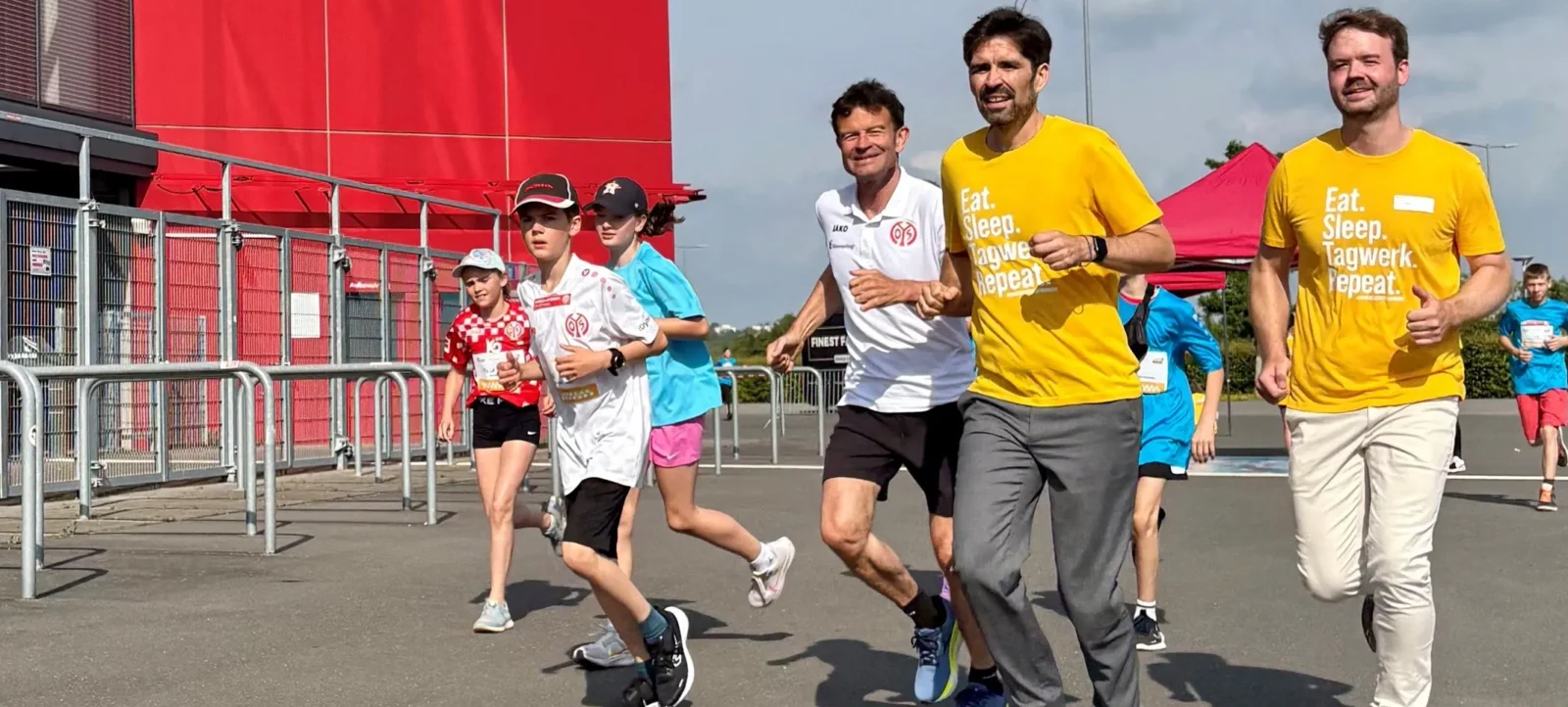 Eine Gruppe von Menschen, darunter Kinder und Erwachsene, läuft an einem roten Gebäude vorbei. Die Läufer tragen verschiedene T-Shirts, einige mit Aufschriften wie 'Eat. Sleep. Tagwerk. Repeat.' und 'Tagwerk'. Die Teilnehmer scheinen an einem Staffellauf oder einer ähnlichen Veranstaltung teilzunehmen, möglicherweise im Rahmen einer Aktion oder eines Events zur Förderung von Gemeinschaft oder Nachhaltigkeit.