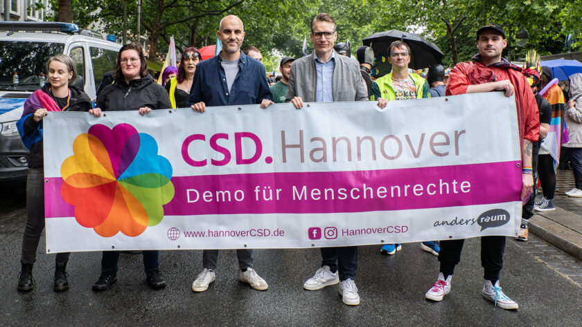 Gruppe von Menschen bei einer Demonstration für Menschenrechte in Hannover. Sie halten ein Banner mit der Aufschrift "CSD Hannover Demo für Menschenrechte" und einem bunten Logo. Die Teilnehmer tragen verschiedene Regenjacken und einige halten Schirme. Im Hintergrund sind weitere Demonstranten und ein Fahrzeug zu sehen.