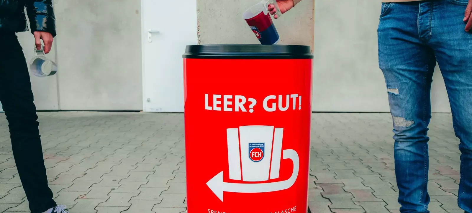 Zwei Personen werfen leere Getränkebehälter in einen roten Müllcontainer mit der Aufschrift 'LEER? GUT!' und 'Spende hier deine Flasche oder deinen Becher'. Der Container hat ein Logo mit den Buchstaben 'FCH' und einen Pfeil, der nach unten zeigt.