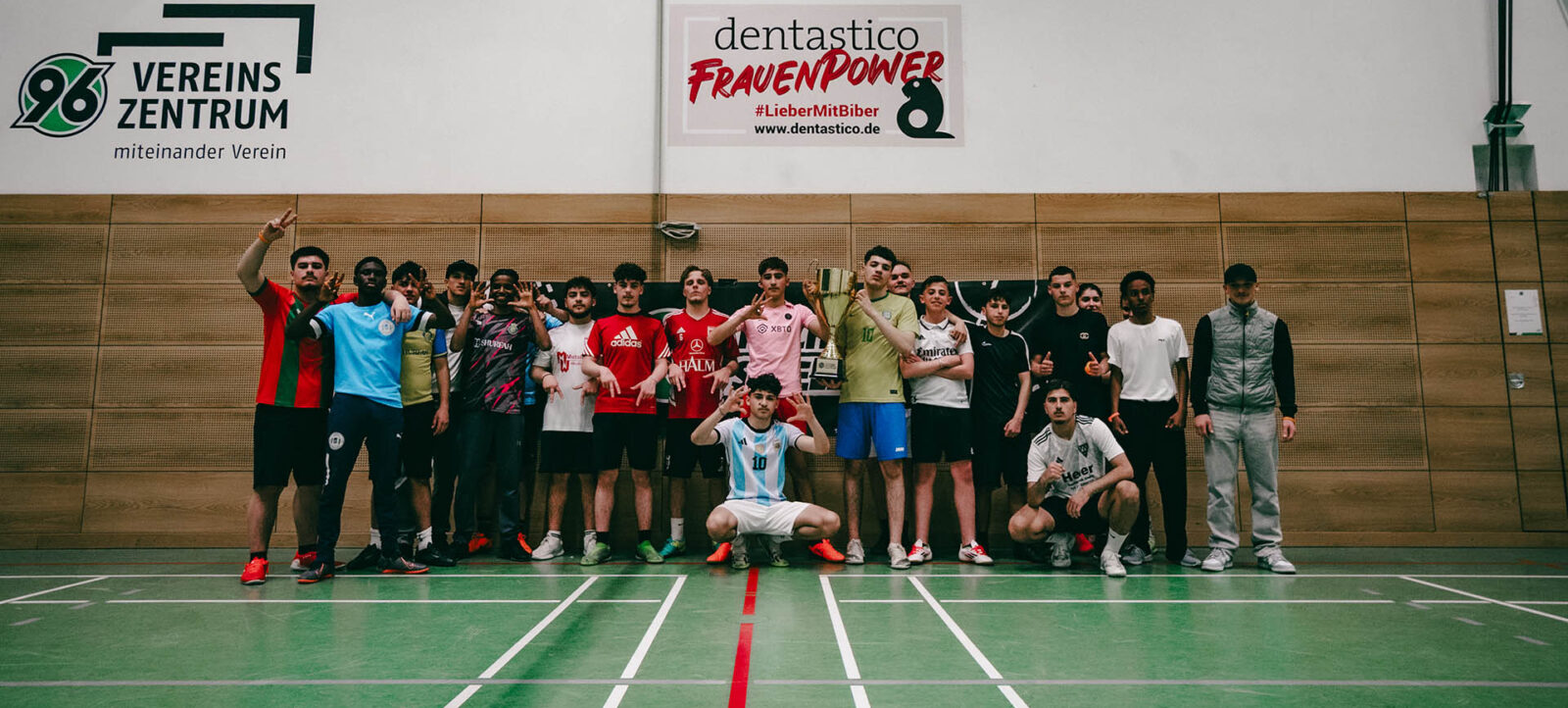 Eine Gruppe von Menschen steht in einer Sporthalle und posiert für ein Gruppenfoto. Sie tragen verschiedene Sportkleidungen und halten einen Pokal. Im Hintergrund sind zwei Schilder zu sehen: eines mit dem Logo und dem Text "Vereins Zentrum miteinander Verein" und ein anderes mit dem Text "dentastico FrauenPower" und einer Hashtag-Kampagne. Die Personen wirken fröhlich und zufrieden, möglicherweise nach einem erfolgreichen Spiel oder Turnier.