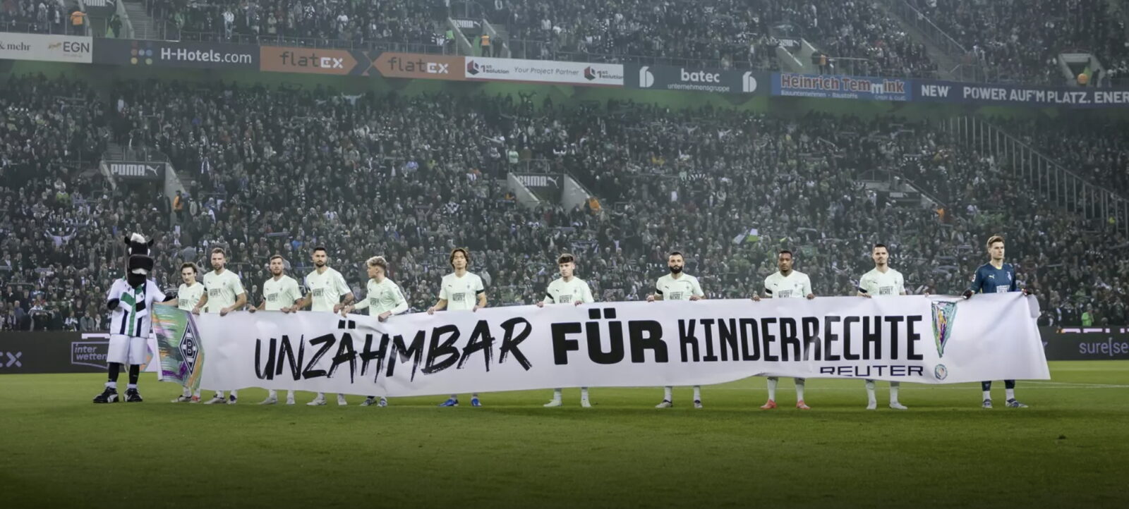 Ein Fußballteam steht auf dem Spielfeld und hält ein großes Transparent mit der Aufschrift "Unzähmbare für Kinderrechte". Im Hintergrund ist eine große Menge von Zuschauern im Stadion zu sehen. Die Spieler tragen grüne Trikots und stehen vor einer Werbebande mit verschiedenen Logos von Sponsoren.