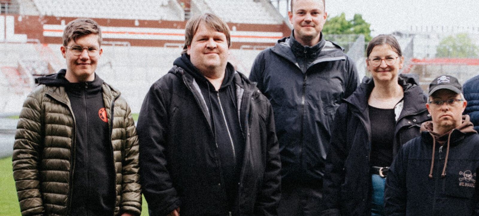 Eine Gruppe von fünf Personen steht auf einem Fußballfeld und lächelt in die Kamera. Im Hintergrund sind die Tribünen des Stadions zu sehen. Die Personen sind in warme Jacken gekleidet, was auf kühleres Wetter hindeutet.