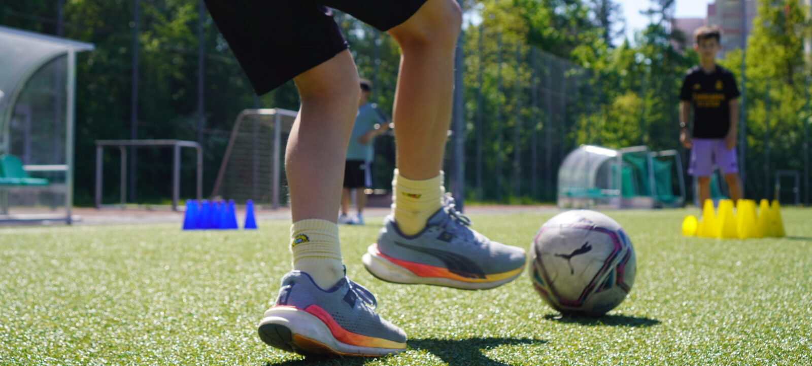 Ein Kind in Sportkleidung steht auf einem Fußballfeld und übt Ballkontrolle mit einem Fußball. Im Hintergrund sind weitere Kinder und Trainingsausrüstung wie Hütchen und Tore zu sehen.