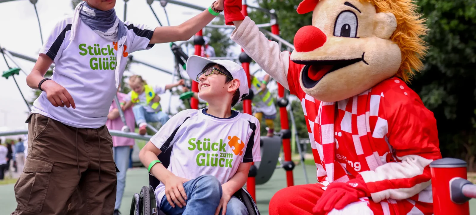Ein Junge im Rollstuhl und ein Begleiter in einem "Stück zum Glück"-T-Shirt stehen auf einem Spielplatz. Ein Clown in einem roten und weißen Kostüm gibt dem Jungen im Rollstuhl einen High-Five. Im Hintergrund sind weitere Kinder und Spielplatzgeräte zu sehen.