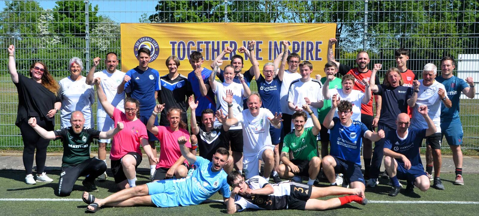Eine Gruppe von Menschen posiert fröhlich auf einem Fußballfeld vor einem Zaun mit einem gelben Banner, auf dem "TOGETHER WE INSPIRE" steht. Die Personen tragen verschiedene Trikots und lächeln oder machen freudige Gesten. Zwei Personen liegen auf dem Boden, während die anderen stehen oder knien.