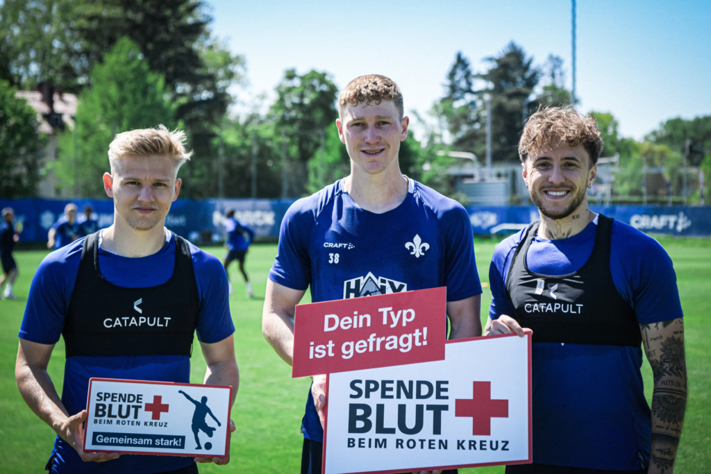 Drei Fußballspieler in blauen Trikots stehen auf einem Fußballfeld und werben für eine Blutspendeaktion. Sie halten Schilder mit den Aufschriften "Dein Typ ist gefragt!" und "Spende Blut beim Roten Kreuz".