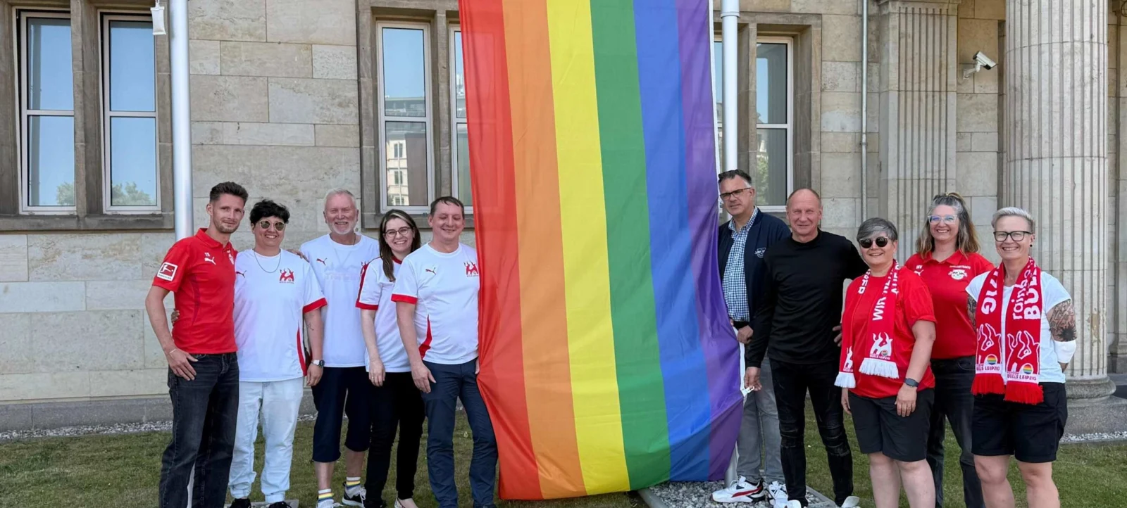 Eine Gruppe von Menschen steht vor einem Gebäude und hält eine große Regenbogenfahne, die für die LGBTQ+-Community steht. Die Personen tragen T-Shirts und Schals mit dem Logo von RB Leipzig, was auf eine Verbindung zu dem Fußballverein hinweist. Sie lächeln und posieren für das Foto, was auf eine unterstützende und inklusive Atmosphäre hinweist.