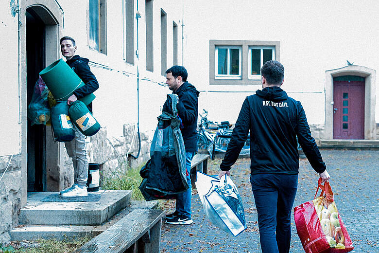Drei Männer tragen verschiedene Gegenstände, darunter Matratzen, Taschen und Einkaufsbeutel, vor einem Gebäude. Einer der Männer trägt eine Jacke mit der Aufschrift "NSC Tat Gut". Sie scheinen dabei zu sein, Umzugsgegenstände zu transportieren.