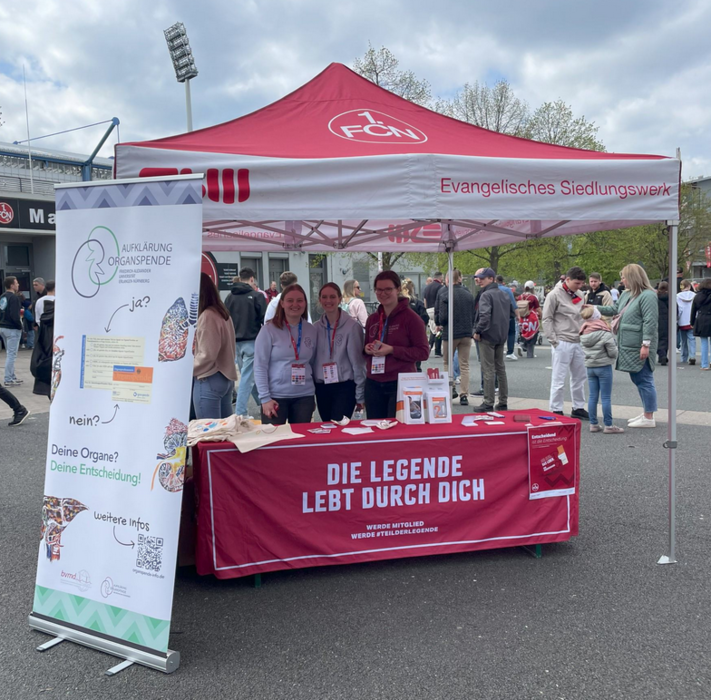 Ein Informationsstand des Evangelischen Siedlungswerks zum Thema Organspende. Drei Personen stehen hinter dem Stand und informieren Besucher über Organspende. Der Stand ist mit einem roten Zelt und einem Banner mit der Aufschrift "Die Legende lebt durch dich" ausgestattet. Ein Plakat mit Informationen zur Organspende ist ebenfalls vorhanden.