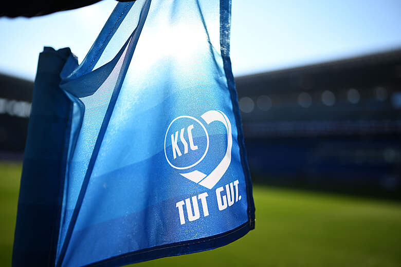 Eine blaue Stofftasche mit dem Logo des Karlsruher SC und dem Slogan "TUT GUT." hängt an einem Geländer vor einem Fußballstadion.