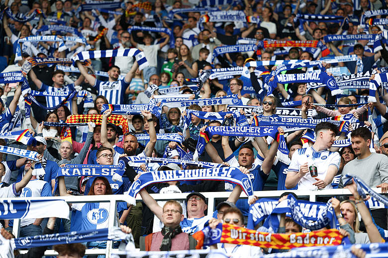 Eine große Gruppe von Fußballfans im Stadion, die alle blaue und weiße Schals und Trikots tragen. Die Fans sind begeistert und halten Schals und Banner hoch, auf denen "Karlsruher SC" und "Meine Heimat" zu lesen ist. Die Atmosphäre ist lebhaft und voller Unterstützung für ihr Team.