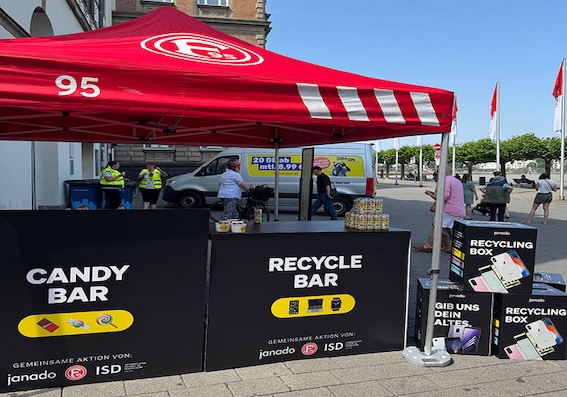 Ein roter Pavillon mit der Aufschrift "95" und dem Logo des 1. FC Köln steht auf einem öffentlichen Platz. Darunter befinden sich zwei schwarze Tische mit den Aufschriften "CANDY BAR" und "RECYCLE BAR". Auf dem "CANDY BAR"-Tisch sind verschiedene Süßigkeiten und Snacks zu sehen, während der "RECYCLE BAR"-Tisch Informationen über das Recycling von Altgeräten bietet. Im Hintergrund sind Menschen zu sehen, die an dem Stand vorbeigehen oder sich dort aufhalten.
