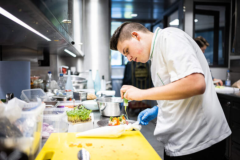 Ein Koch in weißer Kleidung und blauen Handschuhen bereitet in einer modernen Küche ein Gericht zu. Er konzentriert sich auf das Arrangieren von Zutaten auf einem Teller, während verschiedene Küchengeräte und frische Kräuter im Hintergrund zu sehen sind.