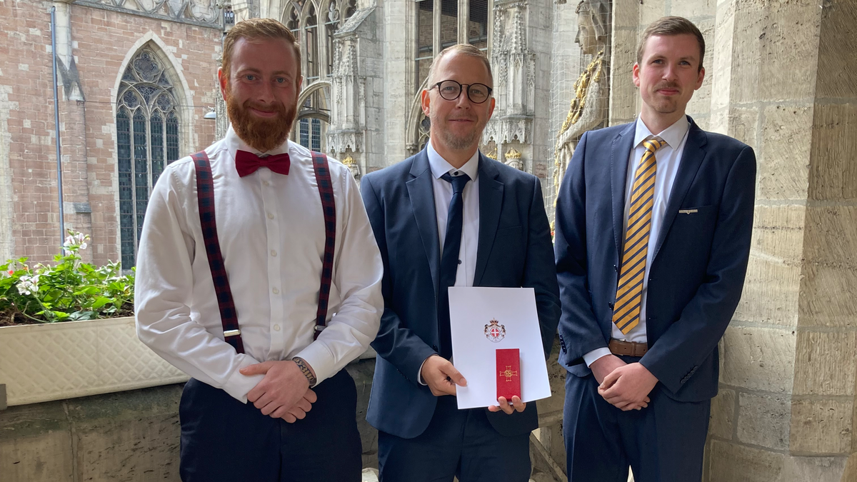 Drei Männer in Anzug stehen vor einem historischen Gebäude mit gotischen Fenstern. Der Mann in der Mitte hält eine Urkunde mit einem Wappen. Die Männer lächeln und scheinen sich über eine Auszeichnung zu freuen.