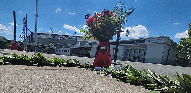 Ein großer Blumenstrauß steht vor einem Stadion. Im Hintergrund sind Teile des Stadions und ein klarer blauer Himmel zu sehen.