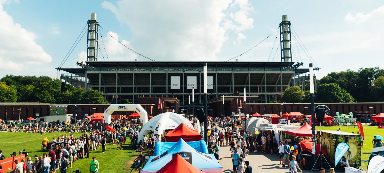 Ein belebter Event vor einem großen Stadion. Menschen strömen zu verschiedenen Zelten und Ständen, die auf dem Rasen aufgestellt sind. Im Vordergrund sind bunte Zelte mit Logos von Sponsoren zu sehen, darunter ein großes rotes Zelt mit blauen und weißen Akzenten. Im Hintergrund steht das Stadion mit zwei markanten Türmen und dem Schriftzug "Rhein Energie Stadion". Die Atmosphäre ist lebhaft und sonnig, mit vielen Besuchern, die an verschiedenen Aktivitäten teilnehmen.