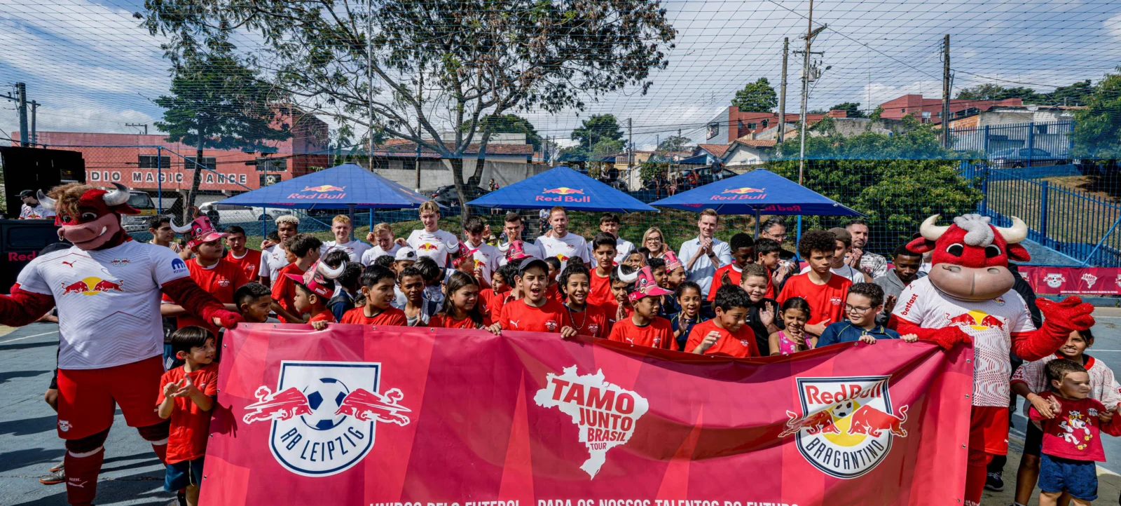 Eine Gruppe von Kindern und Erwachsenen steht vor einem Fußballplatz und hält ein rotes Banner mit den Logos von RB Leipzig und Red Bull Bragantino. Zwei Maskottchen in roten Kostümen mit Stierköpfen stehen links und rechts neben der Gruppe. Im Hintergrund sind blaue Zelte mit dem Red Bull-Logo zu sehen. Die Atmosphäre wirkt fröhlich und gesellig, und die Gruppe scheint sich auf ein gemeinsames Fußballereignis oder eine Veranstaltung vorzubereiten.