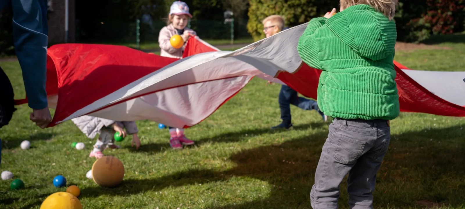 Ein Kind in einem grünen Pullover hält ein großes rotes und weißes Tuch, während es auf einem Rasen steht, auf dem bunte Bälle verstreut sind. Im Hintergrund sind weitere Kinder und ein Erwachsener zu sehen, die an einer Aktivität teilnehmen.