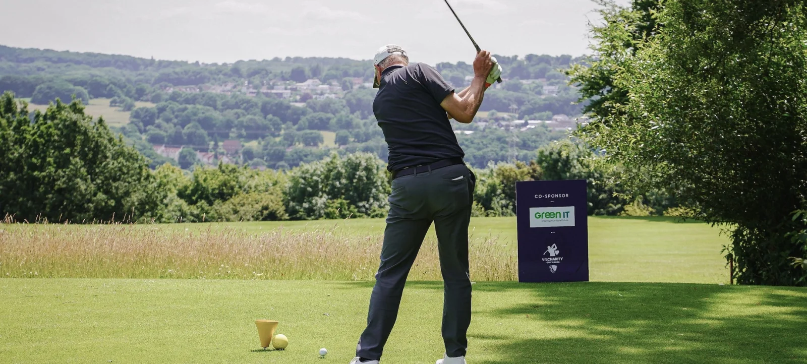 Ein Golfer schwingt seinen Schläger auf einem Golfplatz. Im Hintergrund sind grüne Hügel und Bäume zu sehen. Ein Schild mit den Logos der Sponsoren "Green IT" und "Vilcharity" steht auf dem Rasen.