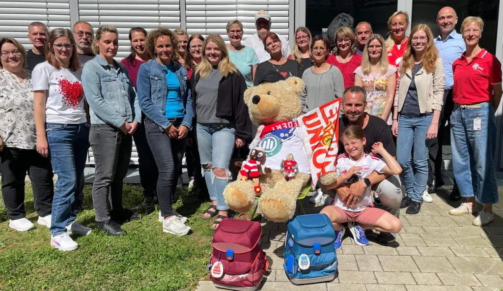 Eine Gruppe von Menschen steht vor einem Gebäude und lächelt in die Kamera. In der Mitte halten zwei Personen einen großen Teddybären und ein Schild mit der Aufschrift "EINHEIT". Vor ihnen auf dem Boden stehen zwei bunte Schulranzen. Die Gruppe scheint sich über etwas zu freuen, möglicherweise im Zusammenhang mit einer Schul- oder Gemeinschaftsaktivität.