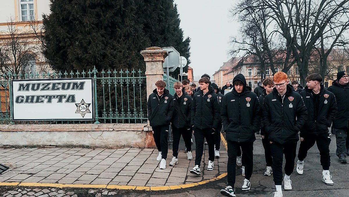Eine Gruppe von Jugendlichen in schwarzen Jacken mit einem Logo auf der Brust geht vor dem "Muzeum Ghetto". Sie bewegen sich auf einem gepflasterten Weg, der von einem schmiedeeisernen Zaun und einem Gebäude mit kahlen Bäumen im Hintergrund umgeben ist.