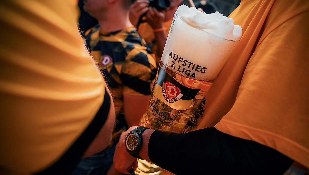 Eine Person hält ein Bierglas mit dem Aufdruck "Aufstieg 2. Liga" und einem DPD-Logo in der Hand. Das Bierglas ist mit weißem Schaum gefüllt. Die Person trägt ein gelbes T-Shirt und eine schwarze Uhr am Handgelenk. Im Hintergrund sind weitere Personen zu sehen, eine davon mit einer Kamera.