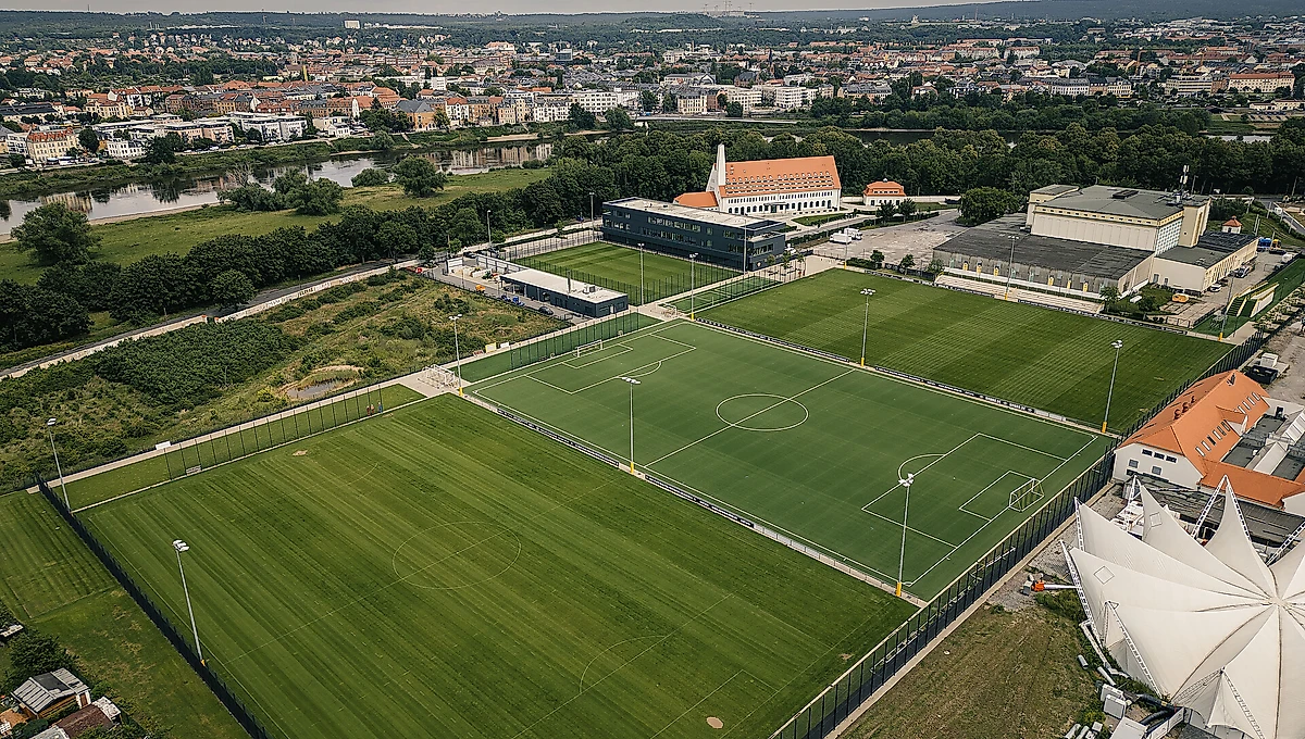 Luftaufnahme eines Fußballplatzes mit Trainingsfeldern und umliegenden Gebäuden. Im Hintergrund ist eine Stadtlandschaft mit Häusern, Grünflächen und einem Fluss zu sehen.