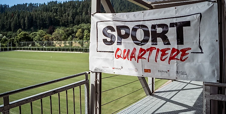 Ein Banner mit der Aufschrift "SPORT QUARTIERE" hängt an einer Tribüne über einem Fußballfeld. Im Hintergrund sind grüne Hügel und Bäume zu sehen.