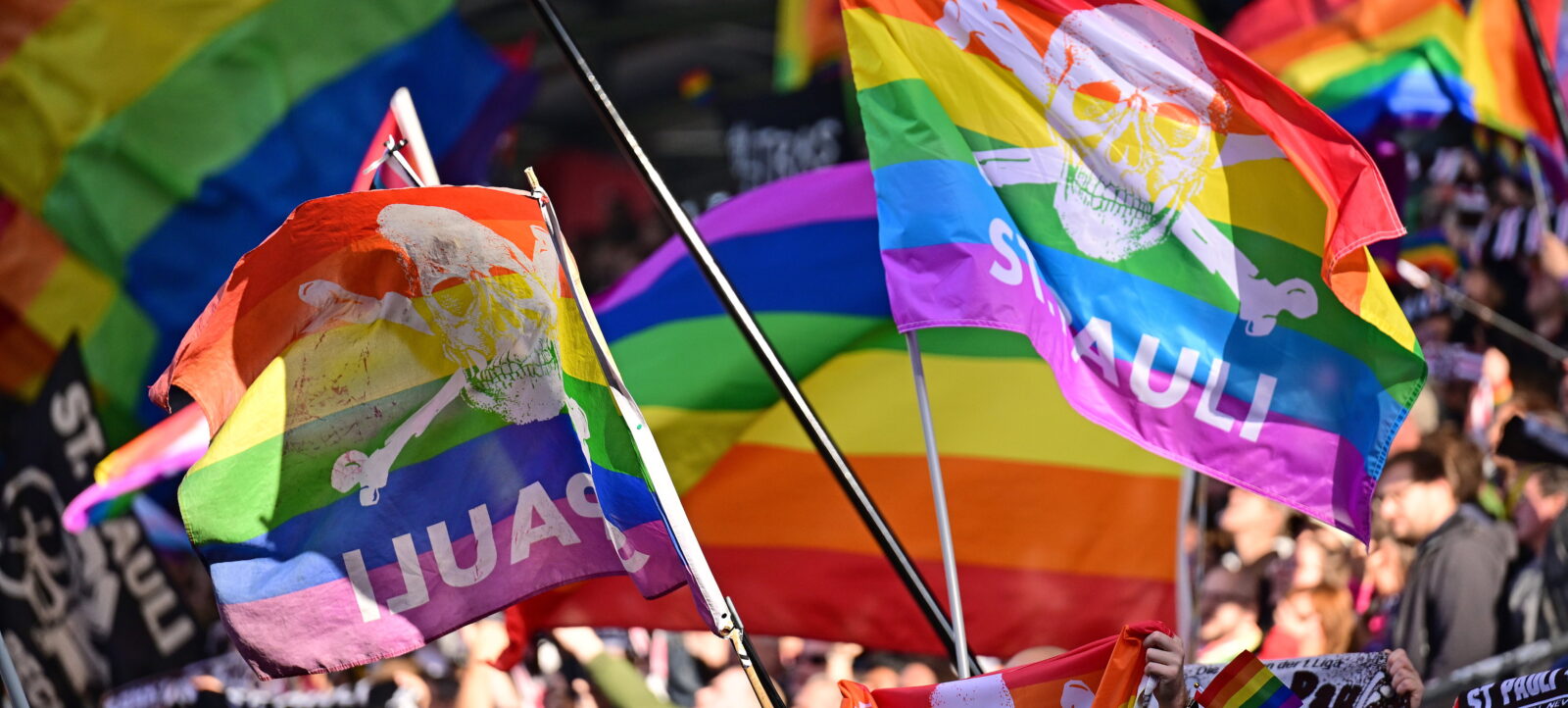 Fans des FC St. Pauli halten Regenbogen-Fahnen mit einem Schädel und den Aufschriften "HUAS" und "JULI" in die Höhe. Die Szene spielt während eines Bundesliga-Spiels im Stadion.