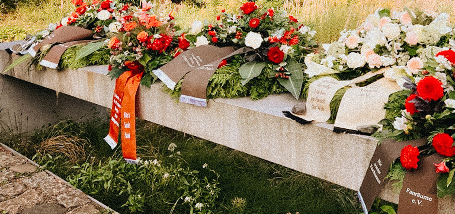 Blumenkränze auf einem Grabstein, umgeben von grünem Gras und Büschen. Die Kränze sind mit roten und weißen Blumen geschmückt und tragen Trauerschleifen mit Aufschriften.