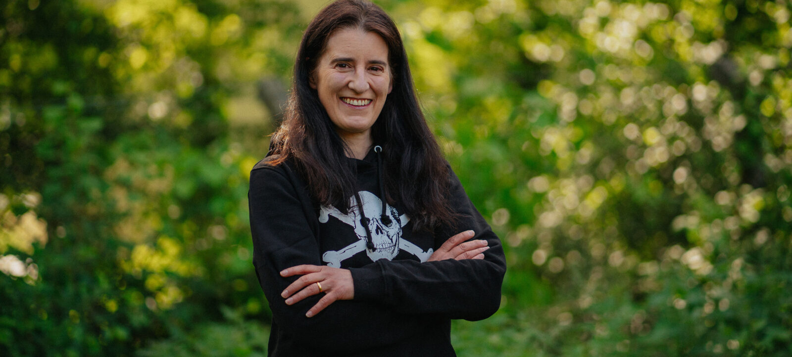 Eine lächelnde Frau mit langen dunklen Haaren steht vor einem grünen, blühenden Hintergrund. Sie trägt ein schwarzes Sweatshirt mit einem weißen Aufdruck, der Knochen und einen Schädel zeigt. Ihre Arme sind vor der Brust verschränkt.