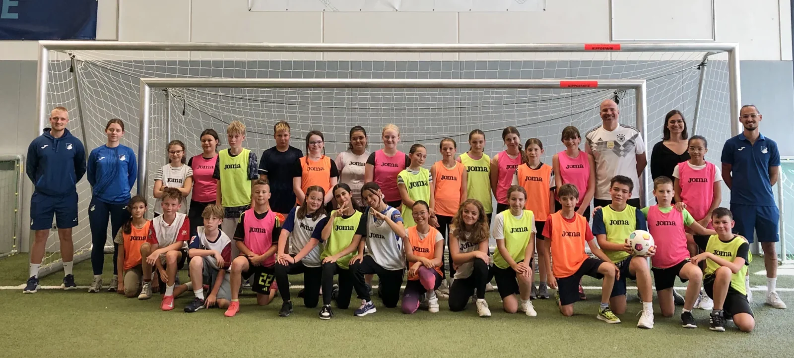 Eine Gruppe von Kindern und Erwachsenen steht vor einem Fußballtor in einer Sporthalle. Die Kinder tragen bunte T-Shirts mit dem Aufdruck "Joma" und sind in verschiedenen Posen für ein Gruppenfoto aufgestellt. Die Erwachsenen, vermutlich Trainer oder Betreuer, stehen an den Seiten und lächeln in die Kamera. Der Hintergrund zeigt die Hallenwand mit einem Tor und einer Werbebande.