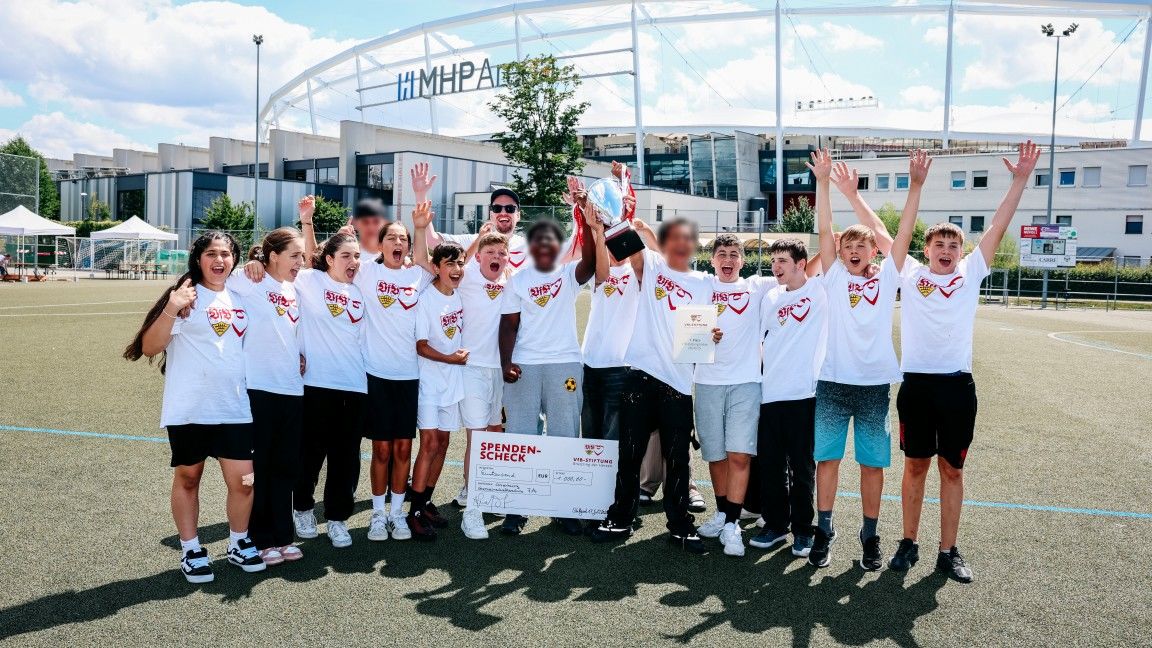 Eine Gruppe junger Menschen steht auf einem Sportplatz vor einem Stadion und feiert einen Erfolg. Sie halten einen Pokal und einen Scheck hoch, während sie fröhlich in die Kamera lächeln. Alle tragen weiße T-Shirts mit einem gemeinsamen Logo und schwarzen Sportkleidung. Der Hintergrund zeigt das Stadion und einige Zelte auf dem Platz.