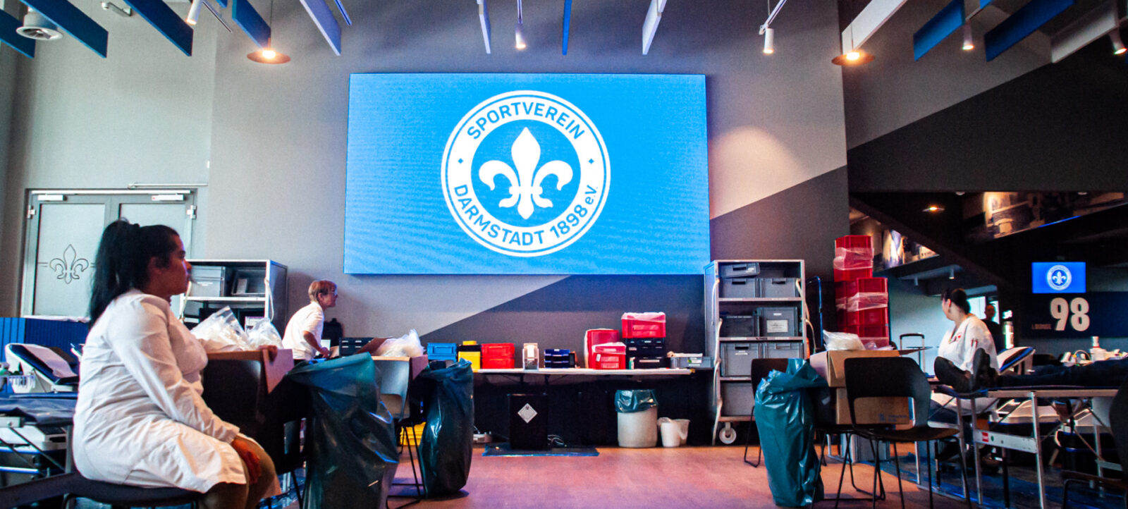 Menschen in einem Raum, die an einer Blutspendeaktion teilnehmen. Im Hintergrund ist ein großes Logo des Sportvereins Darmstadt 1898 zu sehen. Die Teilnehmer sitzen auf Stühlen und sind mit dem Blutspendeprozess beschäftigt. Der Raum ist mit medizinischer Ausrüstung und Müllbeuteln ausgestattet.