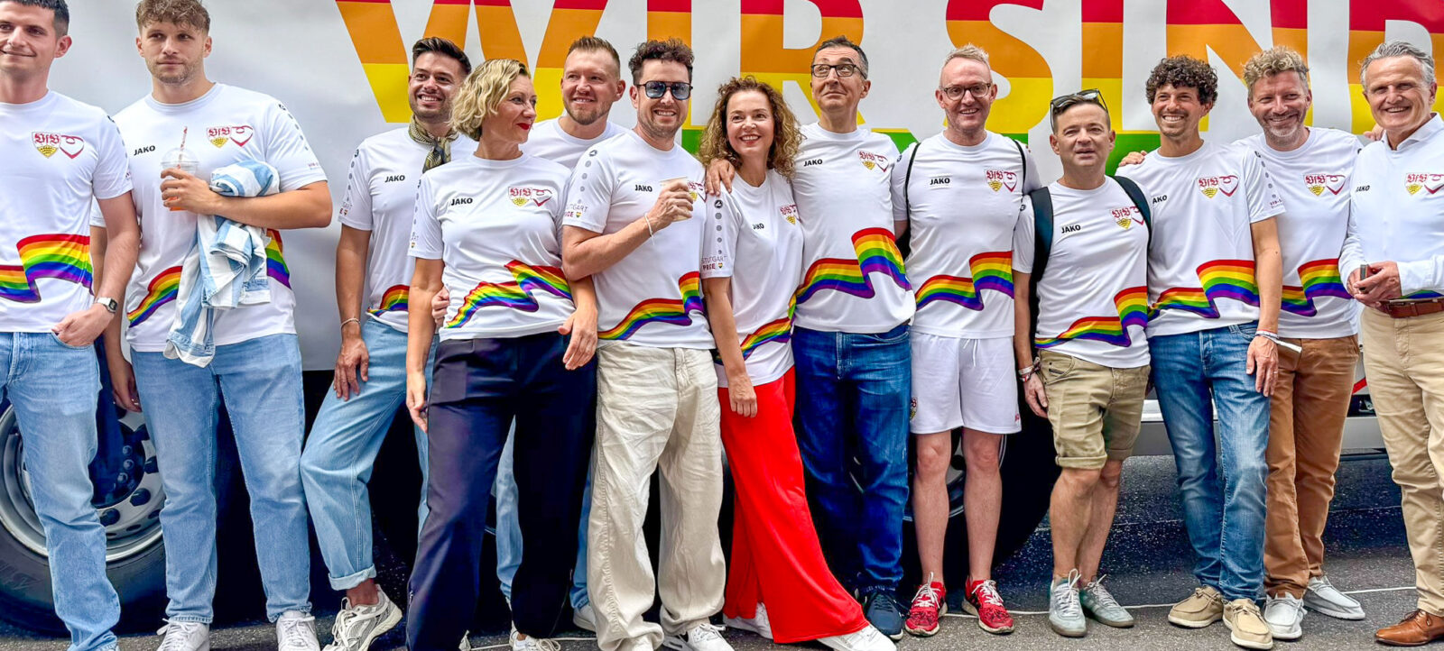 Eine Gruppe von Menschen steht vor einem Banner mit der Aufschrift "WIR SIND". Sie tragen weiße T-Shirts mit einem Regenbogenmuster und dem Logo "JAKO". Die Gruppe lächelt und scheint sich für eine gemeinsame Sache einzusetzen, möglicherweise im Rahmen eines CSD (Christopher Street Day) oder einer ähnlichen Veranstaltung zur Förderung von Vielfalt und Inklusion.