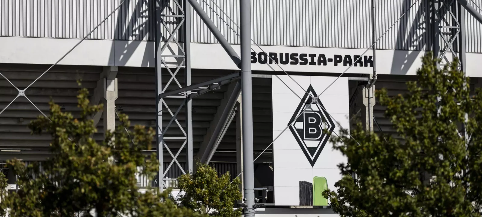 Blick auf das Stadion Borussia-Park mit dem Logo von Borussia Mönchengladbach. Die Struktur des Stadions mit Stahlträgern und der Name 'BORUSSIA-PARK' sind deutlich sichtbar. Im Vordergrund sind Bäume zu sehen.