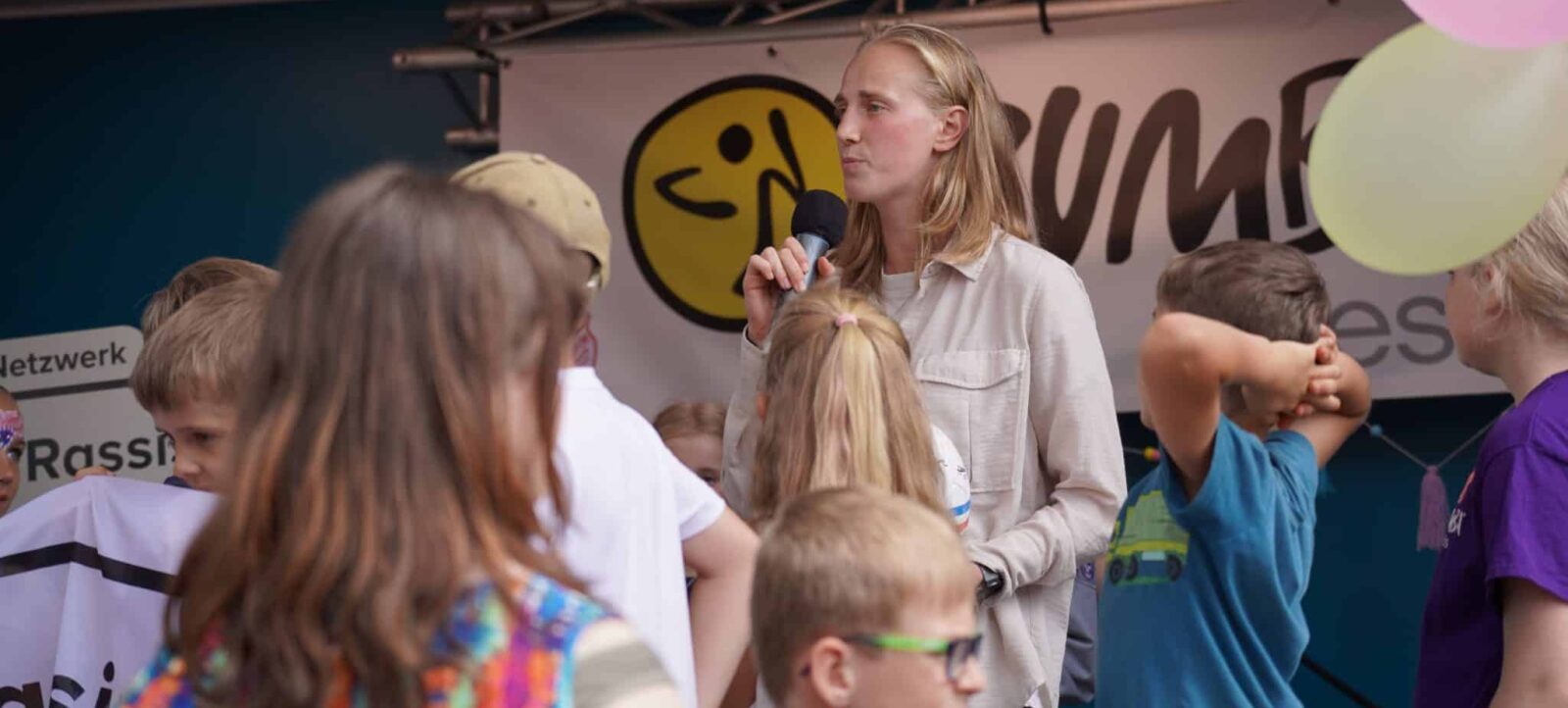 Eine Person mit Mikrofon spricht zu einer Gruppe von Kindern, die aufmerksam zuhören. Im Hintergrund ist ein Banner mit dem Logo und dem Namen eines Netzwerks zu sehen. Die Kinder tragen bunte Kleidung und halten Luftballons.