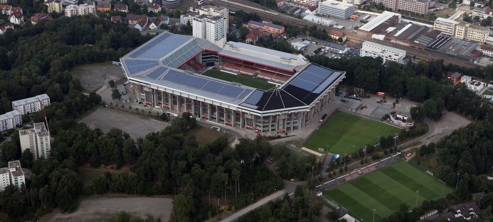 Luftaufnahme des Fritz-Walter-Stadions in Kaiserslautern, umgeben von Grünflächen und Wohngebäuden. Das Stadion ist mit Solarpaneelen auf dem Dach ausgestattet und liegt in der Nähe eines Bahnhofs und weiterer Sportanlagen.