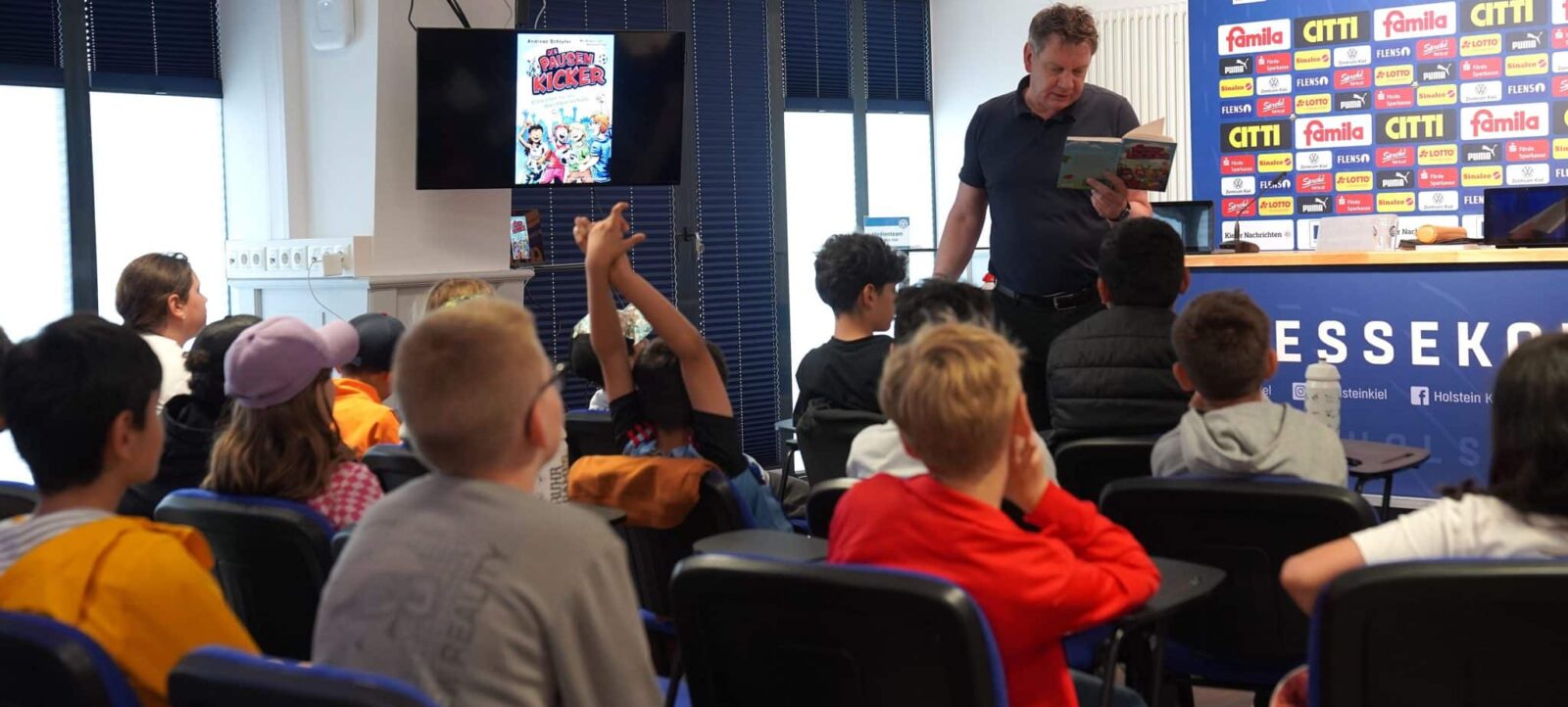 Ein Mann liest Kindern aus dem Buch "Die kleinen Fußball-Kicker" vor. Die Kinder sitzen aufmerksam auf Stühlen und blicken gespannt zum Vorleser. Im Hintergrund sind Werbebanner und ein großer Bildschirm zu sehen, auf dem das Cover des Buches angezeigt wird.