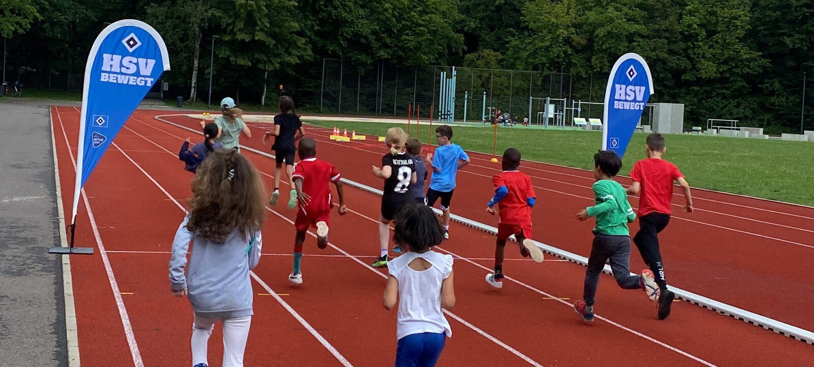Kinder nehmen an einer Sportveranstaltung auf einer Laufbahn teil. Sie laufen in verschiedenen Spuren und scheinen an einem Wettlauf oder einer ähnlichen Aktivität beteiligt zu sein. Im Hintergrund sind zwei blaue Banner mit der Aufschrift "HSV BEWEGT" und dem Logo des HSV zu sehen. Die Veranstaltung findet im Freien auf einer Sportanlage statt, umgeben von Grünflächen und Bäumen.
