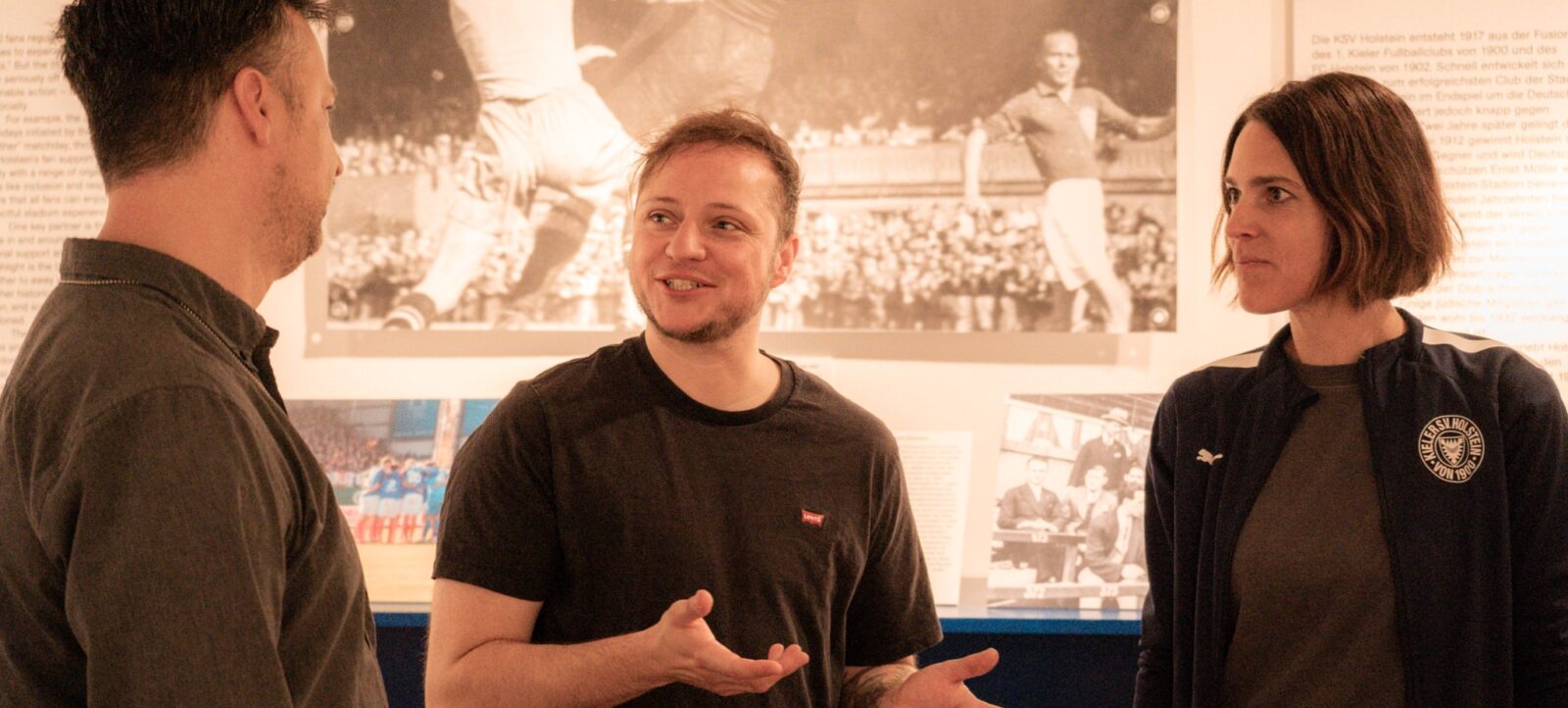 Drei Personen stehen in einer Ausstellung und unterhalten sich. Im Hintergrund sind historische Fotos und Texte zur jüdischen Identität im Fußballstadion zu sehen. Die Personen scheinen sich für die Ausstellung zu interessieren und sind in ein Gespräch vertieft.