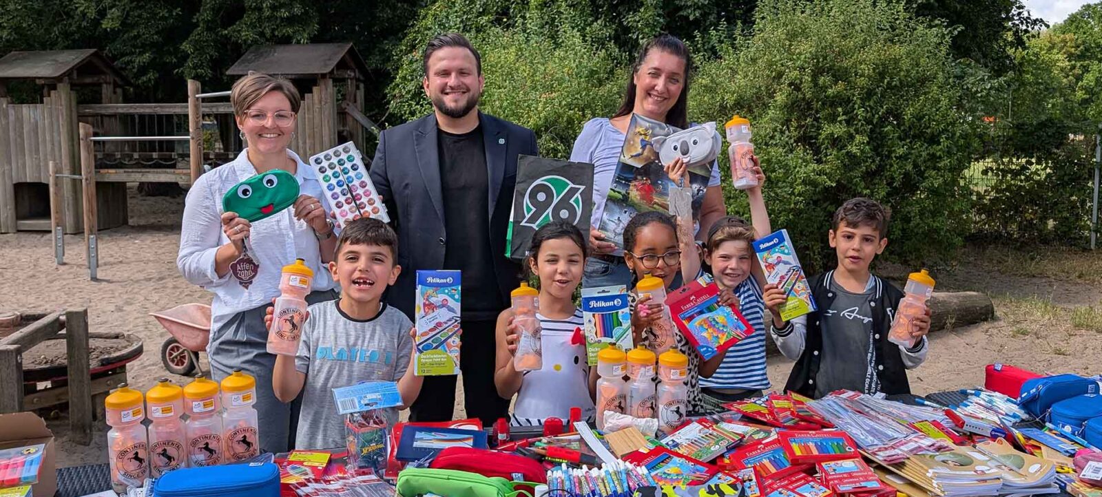 Eine Gruppe von Menschen, darunter Erwachsene und Kinder, steht im Freien und hält verschiedene Schulsachen wie Trinkflaschen, Federmäppchen und Hefte in die Kamera. Vor ihnen liegt ein Tisch voller weiterer Schulsachen. Die Szene findet in einem Park mit Spielgeräten im Hintergrund statt.