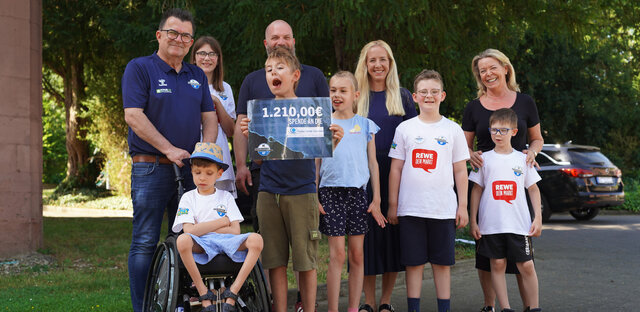 Eine Gruppe von Kindern und Erwachsenen steht vor einem Gebäude und hält ein Schild mit der Aufschrift "1.210.000 Euro für Pauline-Schule". Die Kinder tragen T-Shirts mit dem Logo der Pauline-Schule und einem Vereinslogo. Ein Kind im Rollstuhl ist Teil der Gruppe. Die Atmosphäre ist fröhlich und die Gruppe scheint sich über die erfolgreiche Spendenaktion zu freuen.