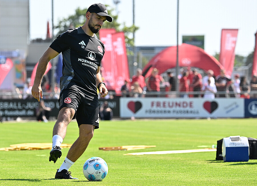 Ein Fußballspieler in schwarzem Trikot und schwarzer Mütze dribbelt einen Fußball auf einem grünen Rasenplatz. Im Hintergrund sind Zuschauer und rote Zelte zu sehen.