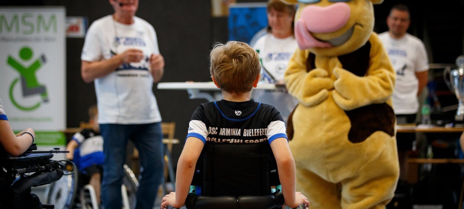 Ein Kind im Rollstuhl mit einem Trikot des DSC Arminia Bielefeld Rollstuhl Sports wird von einer gelben Maske mit Sonnenbrille umarmt. Im Hintergrund stehen Erwachsene in weißen T-Shirts mit dem Logo des MSM (Mobilitäts-Sport-Messe) und ein Plakat mit dem gleichen Logo.