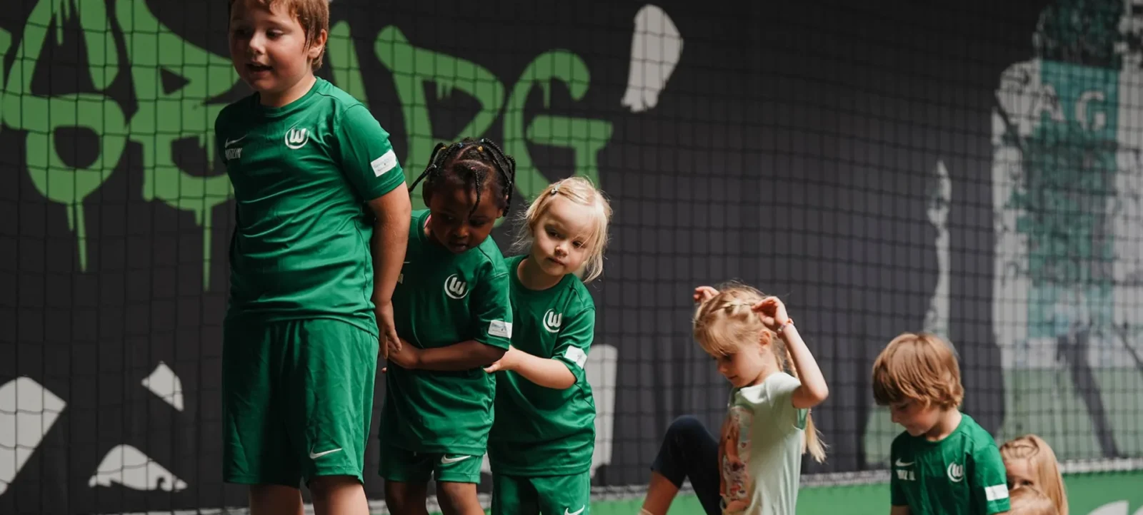 Kinder in grünen Trikots des VfL Wolfsburg nehmen an einem Aktionstag in einer Kita teil. Sie stehen vor einem grünen Netz mit dem Logo des Vereins und dem Text "Kita". Die Kinder wirken konzentriert und engagiert.