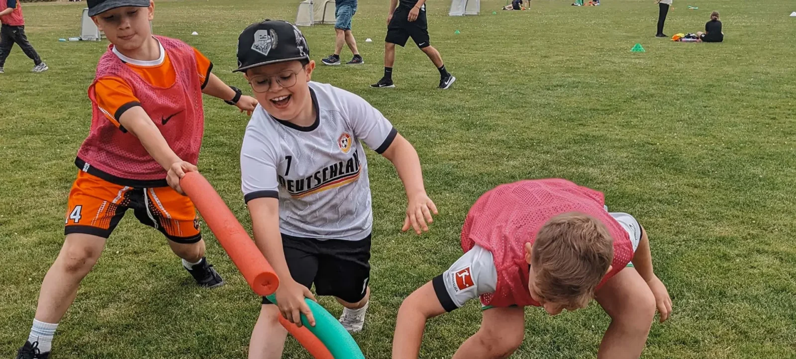 Drei Kinder spielen auf einem Fußballplatz ein Spiel mit bunten Stangen. Die Kinder tragen Sportkleidung und Hüte. Im Hintergrund sind weitere Kinder und Betreuer zu sehen, die an verschiedenen Aktivitäten teilnehmen.