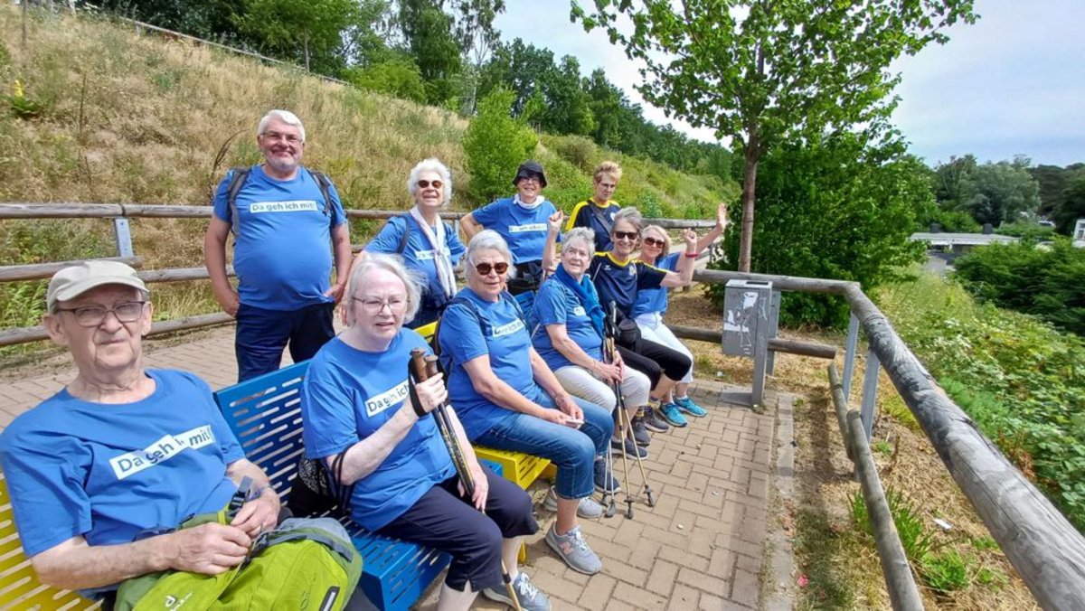 Eine Gruppe älterer Menschen, die auf einer Bank und im Stehen posieren, tragen blaue T-Shirts mit dem Aufdruck "Das geht mich an!" Sie befinden sich in einer grünen, parkähnlichen Umgebung mit Bäumen und einem Bach im Hintergrund.