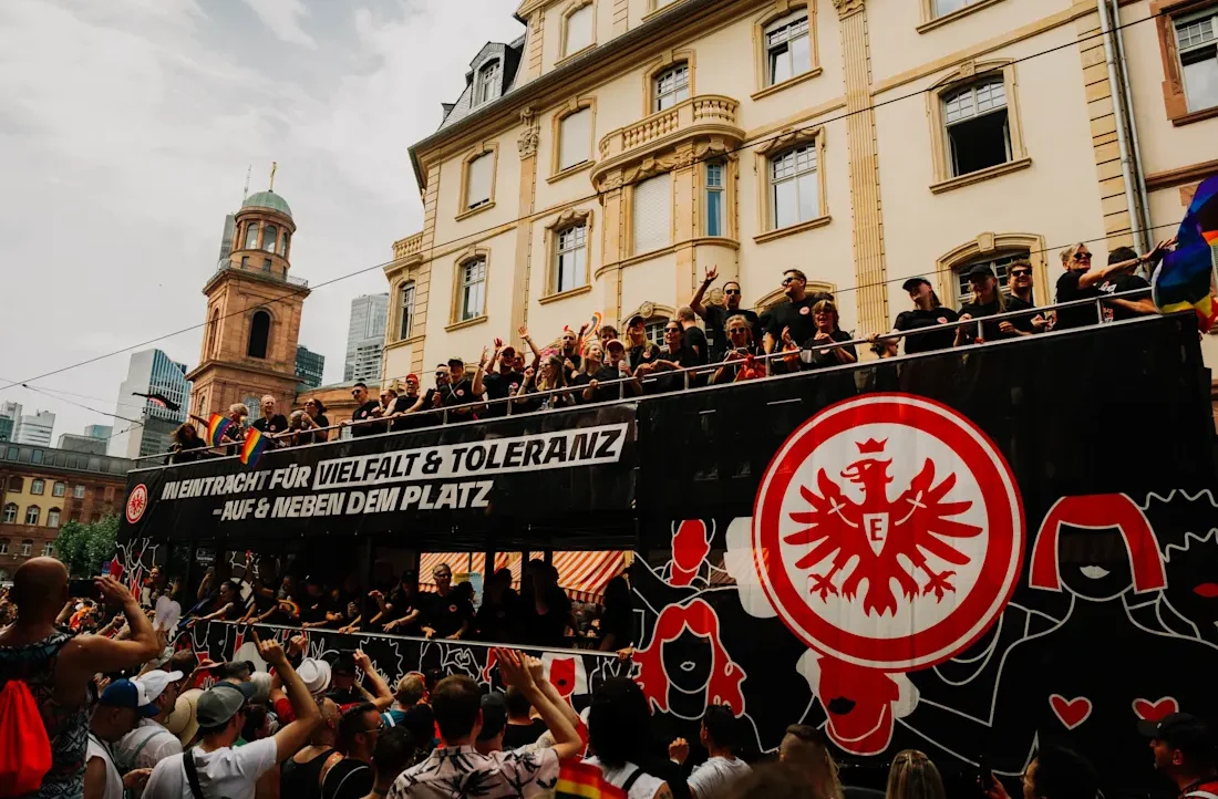 Ein großer, beflaggter Fan-Bus mit dem Logo des 1. FC Köln fährt durch eine Stadt. Auf dem Dach des Busses stehen viele Menschen und halten Fahnen und Schilder hoch. Ein Schild auf dem Bus zeigt die Aufschrift "Eintracht für Vielfalt & Toleranz - Auf & neben dem Platz". Im Hintergrund sind historische Gebäude und eine Kirche zu sehen.