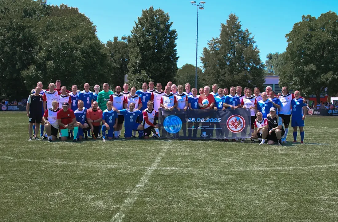 Eine Gruppe von Menschen in Fußballtrikots steht auf einem Rasenfeld für ein Gruppenfoto. Die Trikots sind in verschiedenen Farben, hauptsächlich blau, rot und weiß. Die Gruppe ist in zwei Reihen angeordnet, eine stehend und eine kniend. Sie halten ein Banner mit dem Datum "25.06.2025" und Logos, die auf eine Veranstaltung hinweisen. Im Hintergrund sind Bäume und ein klarer Himmel zu sehen.